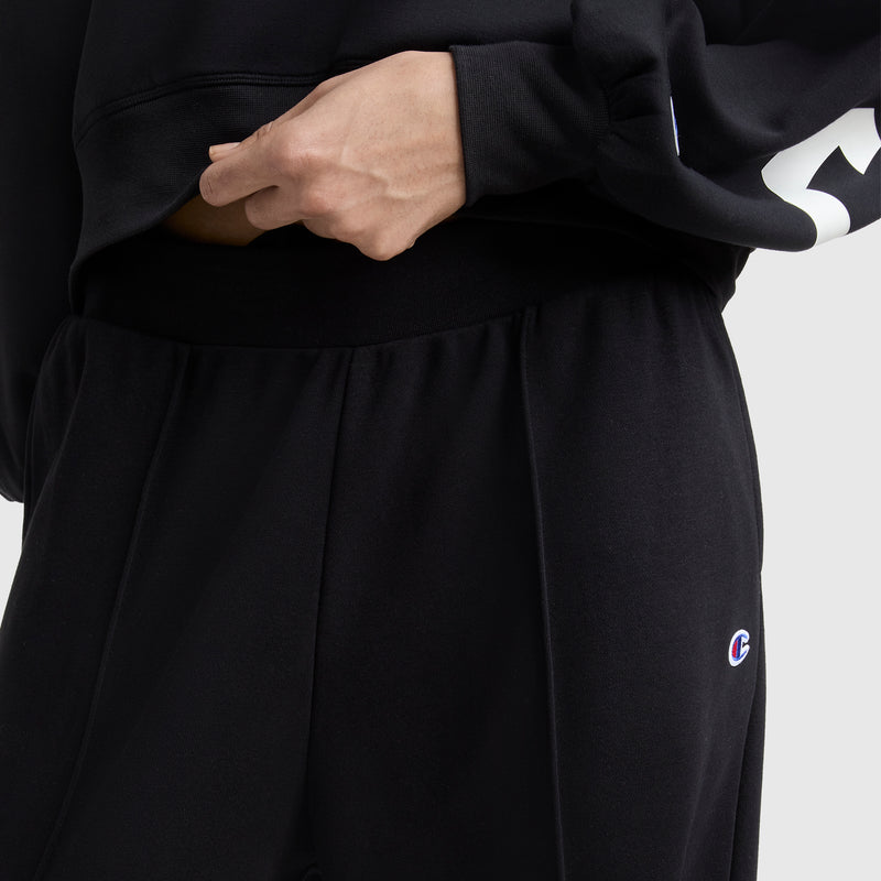 A person is shown from the waist down wearing Champion Tackle Open Bottom Pants with a small C logo, paired with a black French terry sweatshirt, one hand resting on the waistband.
