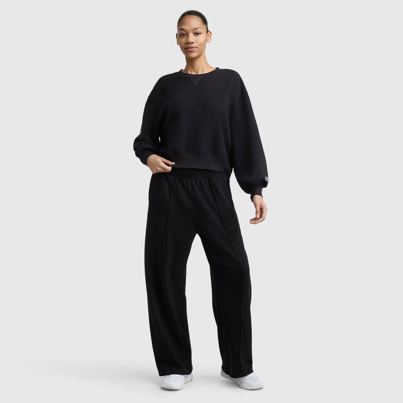 A person stands against a plain light background, wearing a loose black sweatshirt, Champion Tackle Open Bottom Pant with C Logo, and white sneakers.