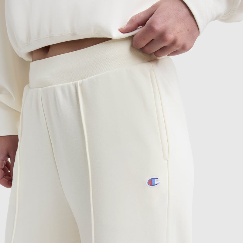 A person wears Champion's Tackle Open Bottom Pant with a C Logo in cream, holding the waistband, paired with a matching cream top.