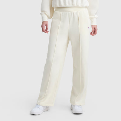 A person wears Champion’s Tackle Open Bottom Pant with C Logo—high-waisted, cream French terry wide-leg pants with front seams—paired with a matching top and white sneakers, standing against a plain light background.