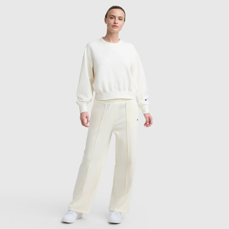 A person stands against a light background wearing Champion's Tackle Open Bottom Pant with C Logo in off-white, paired with a matching French terry sweatshirt and white sneakers, posing casually with one hand relaxed and the other slightly bent.