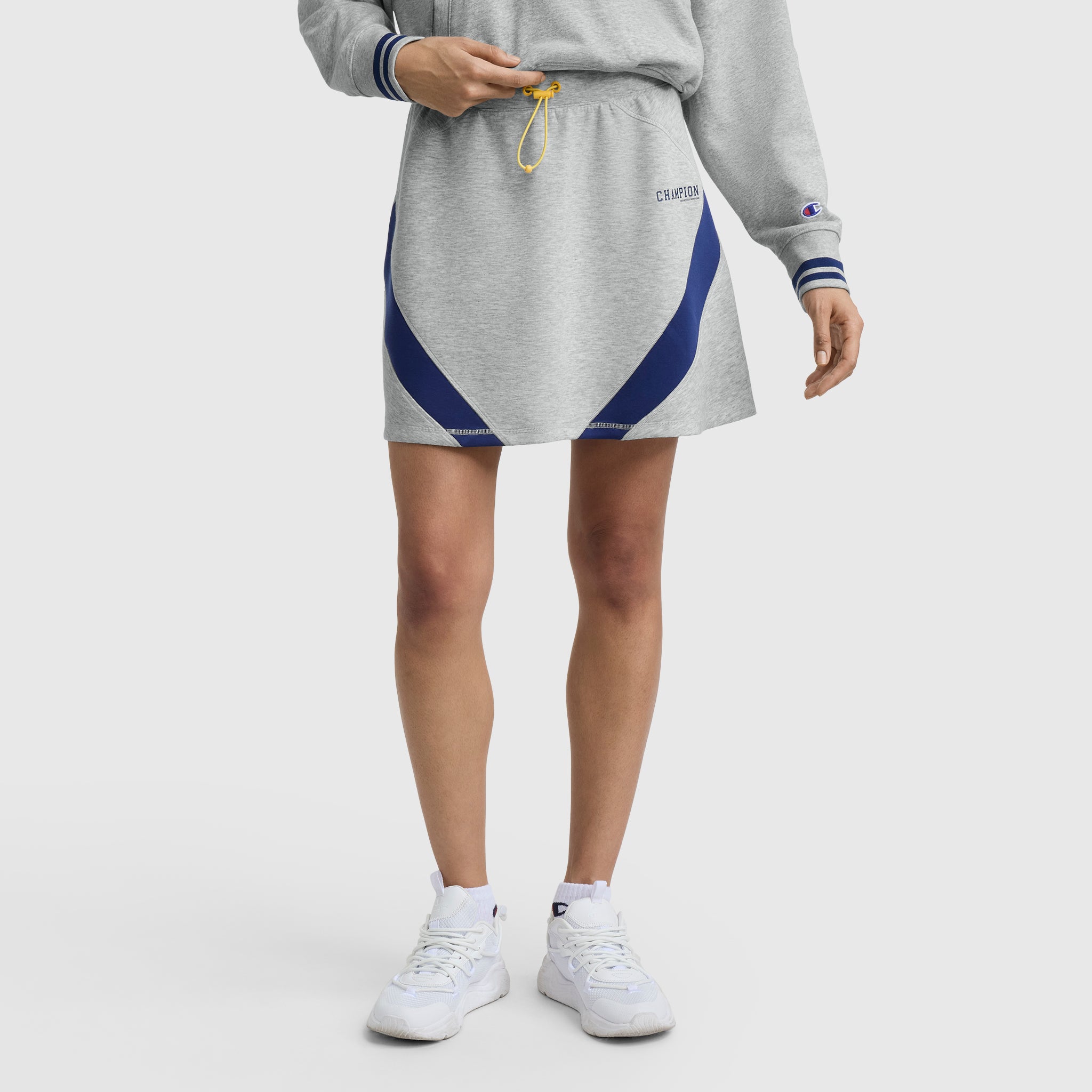 A person wearing a light gray Champion sweatshirt and the Champion Endzone Color Block Skirt with blue accents adjusts the yellow waist drawstring. They also wear white athletic shoes, all against a plain white background.