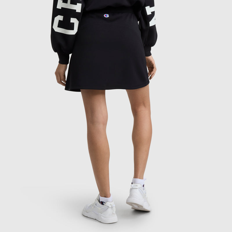 A person stands facing away against a plain light background, wearing a black Champion Endzone Color Block Skirt, a black sweatshirt with white sleeve text, white socks, and white sneakers.