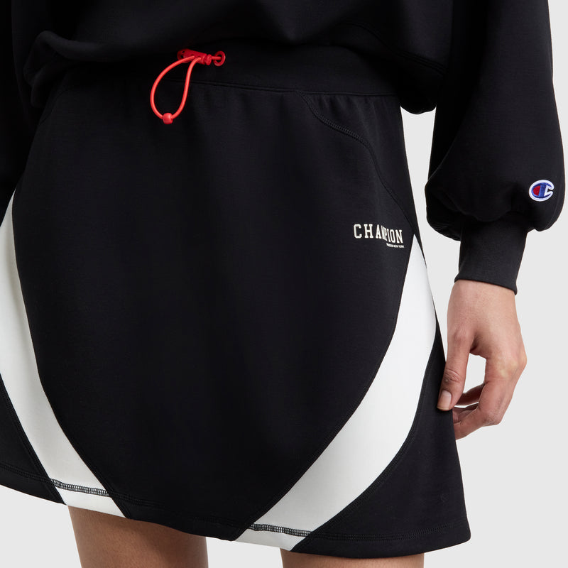 A person wears the Champion Endzone Color Block Skirt featuring white curved panels, a red drawstring, and partial CHAMPION lettering, paired with a black sweatshirt with the Champion logo on the sleeve for a sporty look.