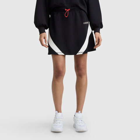 A person wears a Champion Endzone Color Block Skirt with white curved panels, paired with a long-sleeve black Champion sweatshirt and white sneakers, standing against a plain light gray background.