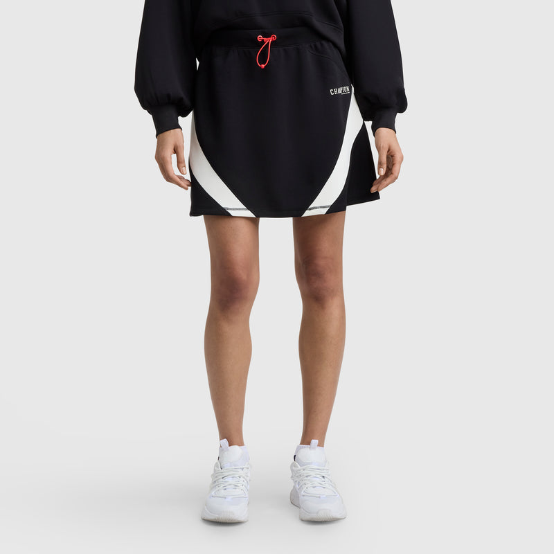 A person wears a Champion Endzone Color Block Skirt with white curved panels, paired with a long-sleeve black Champion sweatshirt and white sneakers, standing against a plain light gray background.