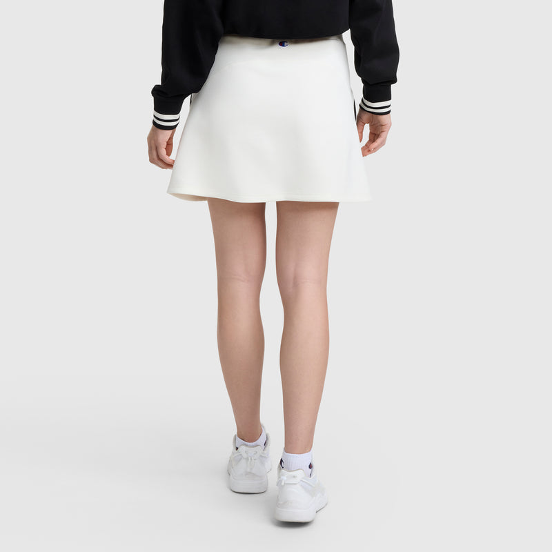 A person faces away, wearing the Champion Endzone Color Block Skirt styled as an athletic skirt, paired with a black Champion jacket with striped cuffs, white sneakers, and socks against a plain light background.