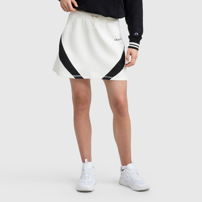 A person wears the Champion Endzone Color Block Skirt—a white skirt with black diagonal stripes—alongside a black long-sleeve top and white sneakers, posing against a plain light gray background.