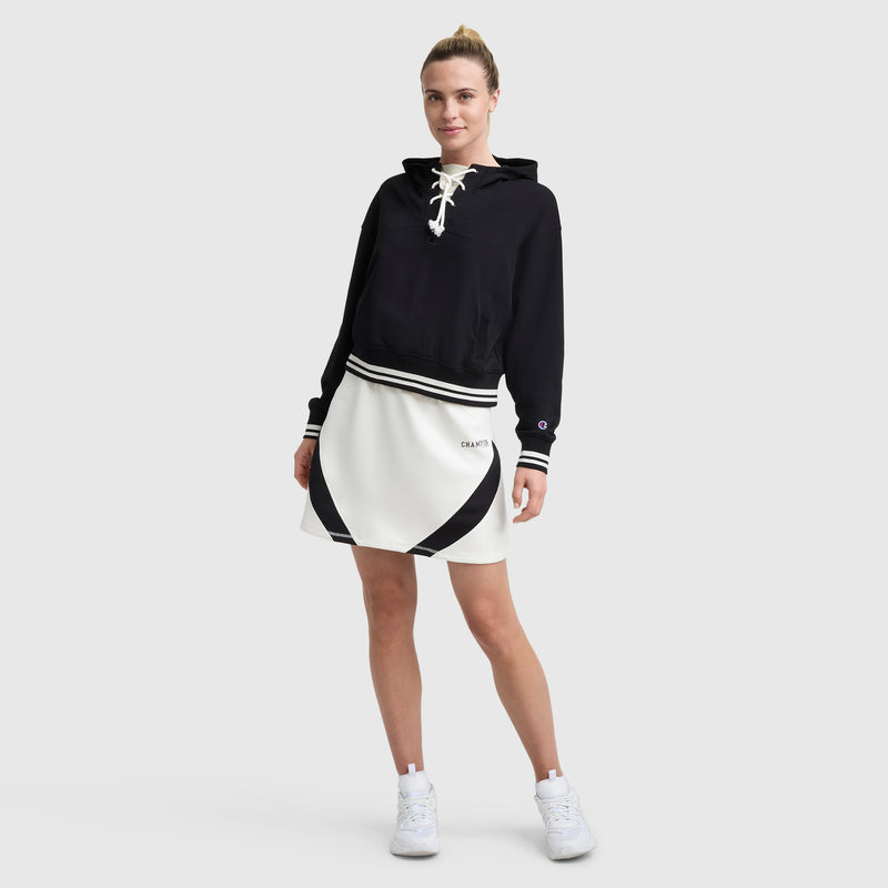 A woman smiles against a light gray background, wearing a black lace-up hoodie, the Champion Endzone Color Block Skirt, and white sneakers. Her hair is tied back.