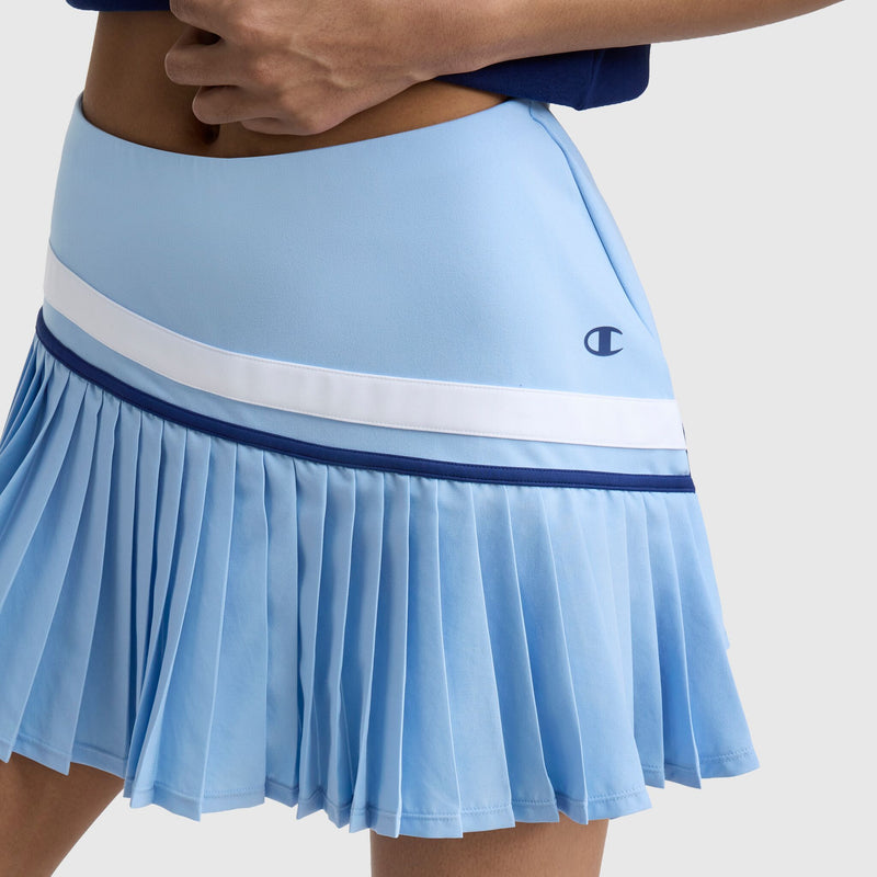 Close-up of a person wearing the Champion Country Club Pleated Skort with white and navy stripes and C logo on the side. The person's hand rests on their waist, and part of a blue top is visible.