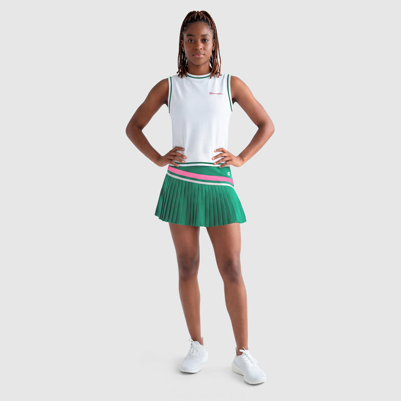 A person stands confidently in a Champion Country Club Pleated Skort with pink and white stripes, made from recycled polyester, paired with a white athletic tank top and sneakers against a light gray background.