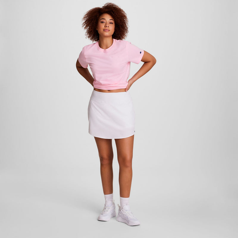 A woman stands with hands on hips wearing a light pink tee, Champion Courtside Skort with C Logo, white socks, and sneakers against a plain white background.