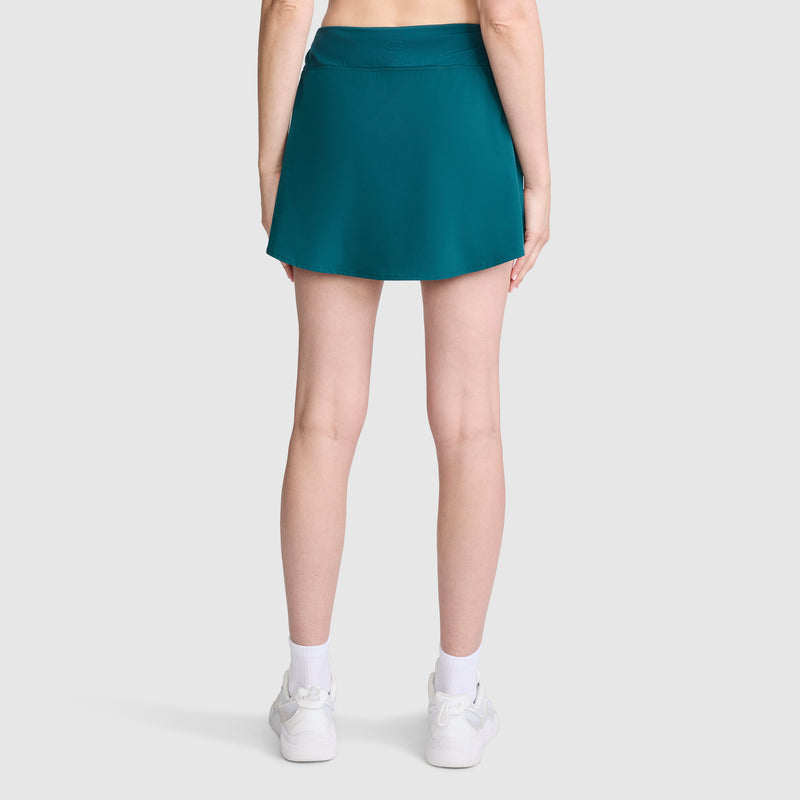 A person stands facing away from the camera against a plain light gray background, wearing the Champion Courtside Skort, C Logo in teal, paired with white socks and white sneakers.