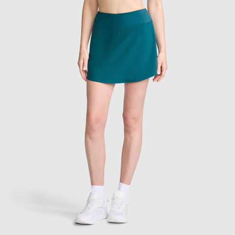 A woman models the Champion Courtside Skort, C Logo in teal, paired with white socks and white sneakers, standing against a plain light gray background. Her upper body and face are not visible in the image.