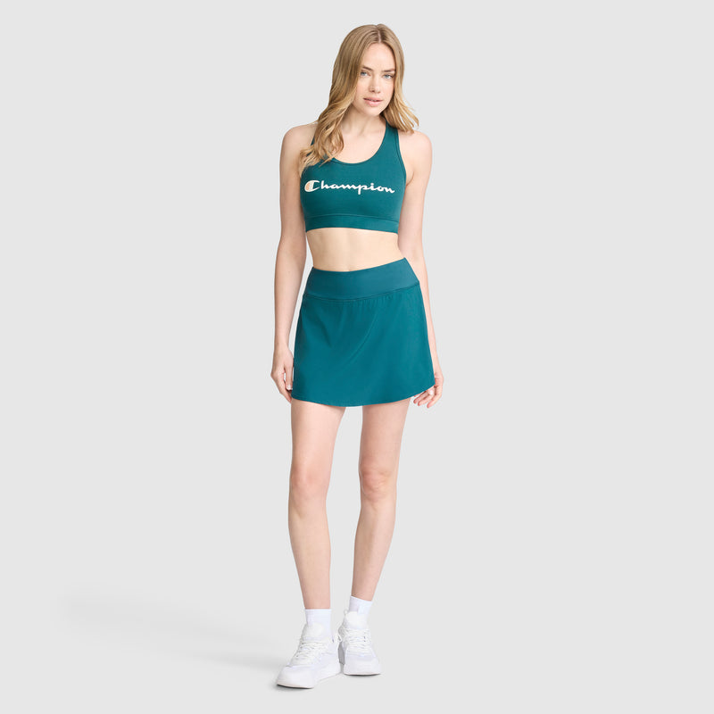 A woman stands against a plain light gray background wearing the Champion Courtside Skort and matching teal C Logo crop top for a fresh court style vibe, paired with white sneakers.