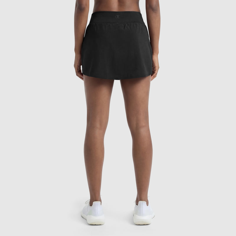 Someone stands with their back to the camera, displaying Champion’s Courtside Skort, C Logo in black along with white athletic shoes against a simple light background. Only the sporty court-inspired lower body is shown.