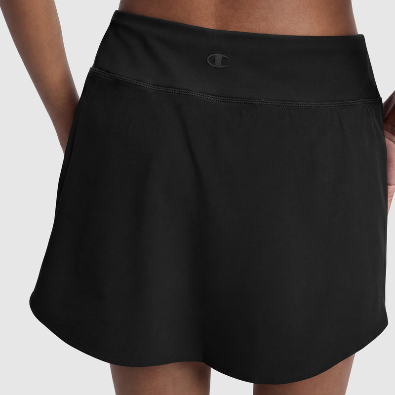 A person wears the Champion Courtside Skort, C Logo, in black with hands in side pockets. The skort fuses court style and sporty function, featuring a wide waistband and a small Champion logo at the center. Plain light gray background.