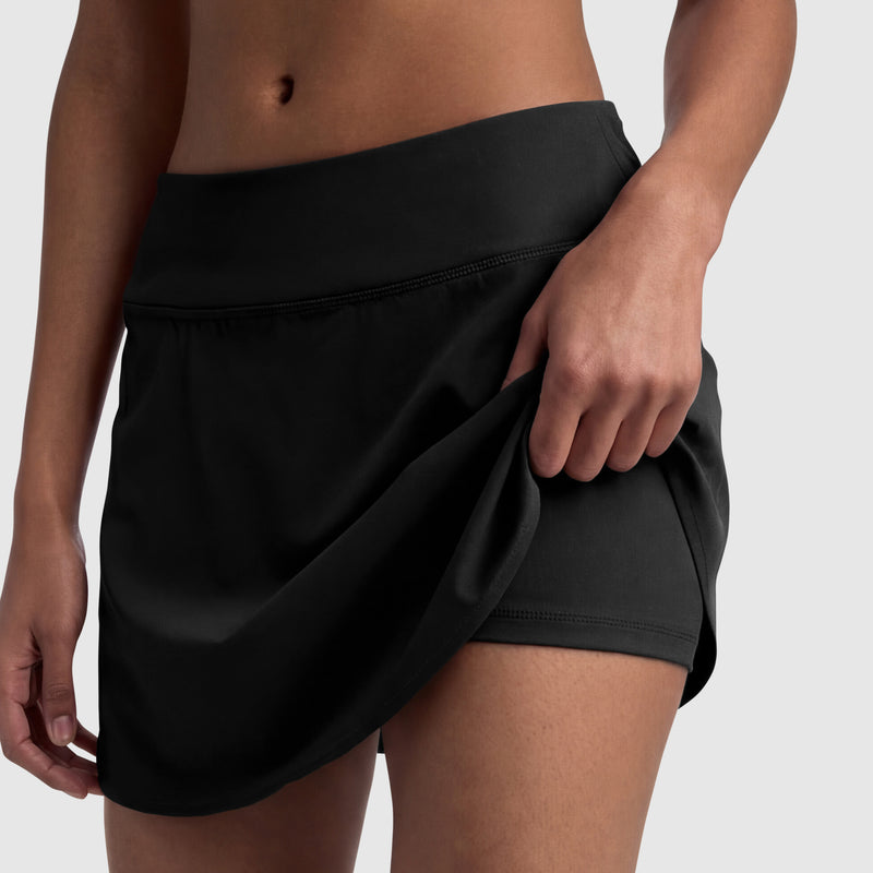 A woman wearing the Champion Courtside Skort with C Logo lifts the hem to reveal fitted shorts underneath, showcasing its sporty design and midsection against a plain background.
