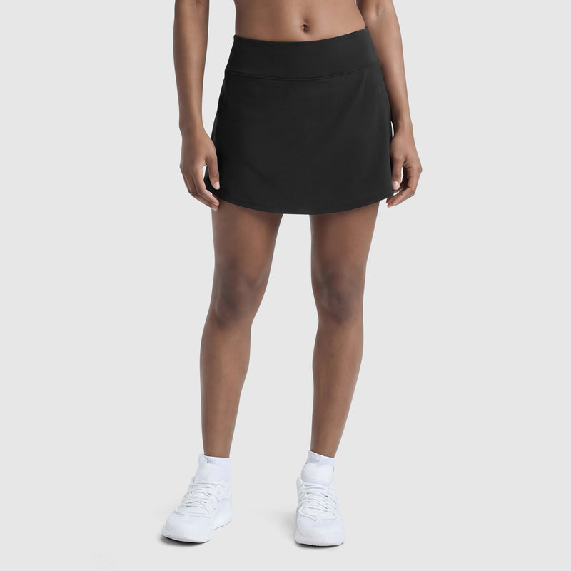 A person models Champion’s Courtside Skort, C Logo in black, styled with white socks and sneakers against a plain light gray background. Only the lower torso and legs are shown.