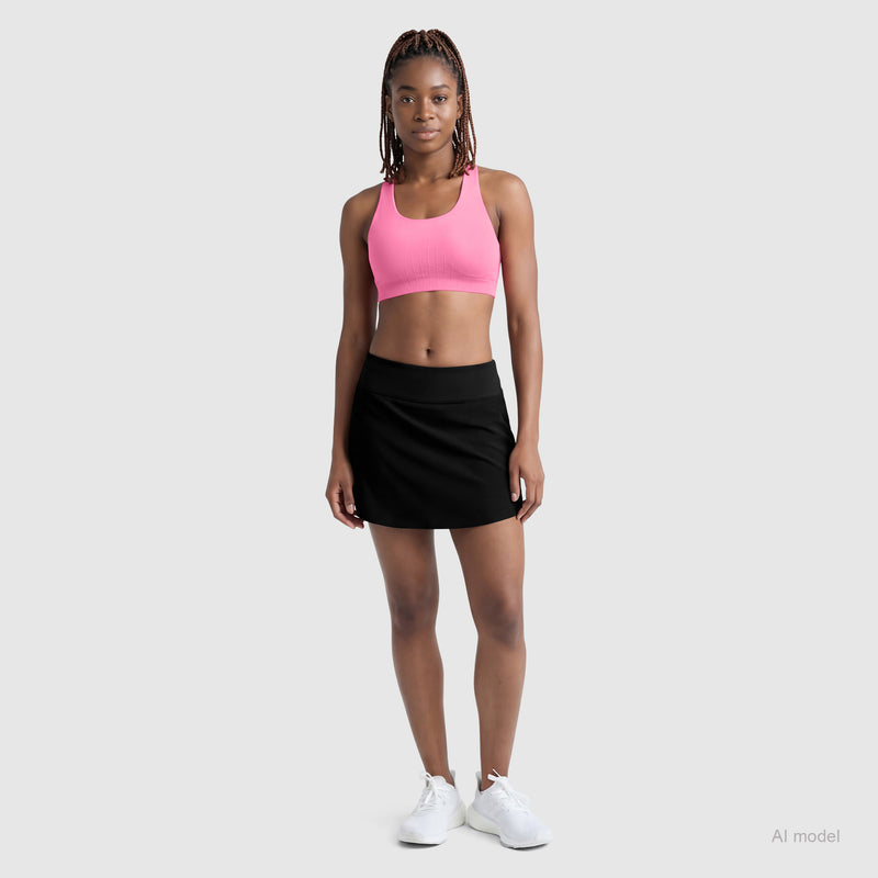 A woman with braided hair and a neutral expression wears the Champion Courtside Skort, a pink sports bra, and white sneakers against a light gray background, showcasing effortless court style with sporty function.