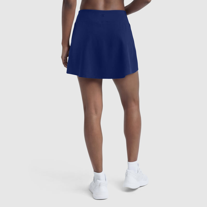 A woman faces away, wearing a Champion Courtside Skort with C Logo and white sneakers. The plain light background highlights the skort's sporty function and court-inspired style.