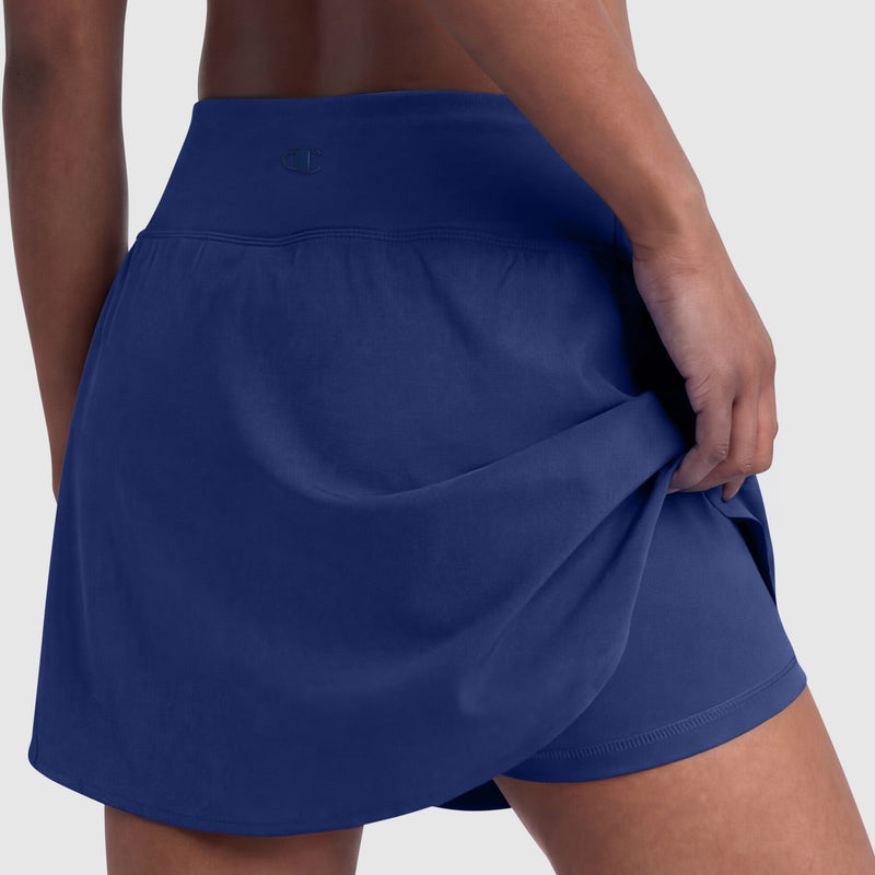 A person in the Champion Courtside Skort, C Logo, navy blue women's skort with attached shorts, lifts the skirt slightly to show its sporty design. The background is plain light gray.