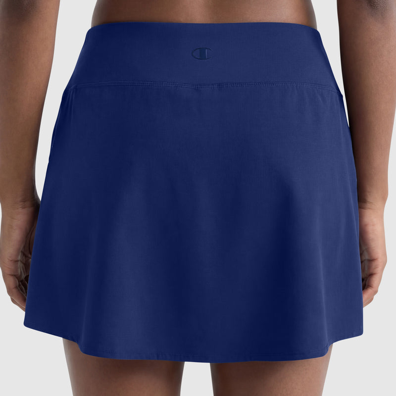 A person is seen from behind wearing the Champion Courtside Skort, C Logo in navy blue. This athletic skirt has an elastic waistband, falls above the knee, and features the Champion logo on the back waistband for court-ready style.