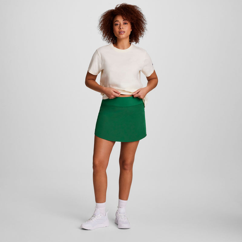 A woman with curly hair wears the Champion Courtside Skort, C Logo, styled with a white t-shirt, white socks, and sneakers. She stands against a plain light gray background, looking forward as she adjusts her shirt for a sporty-casual look.