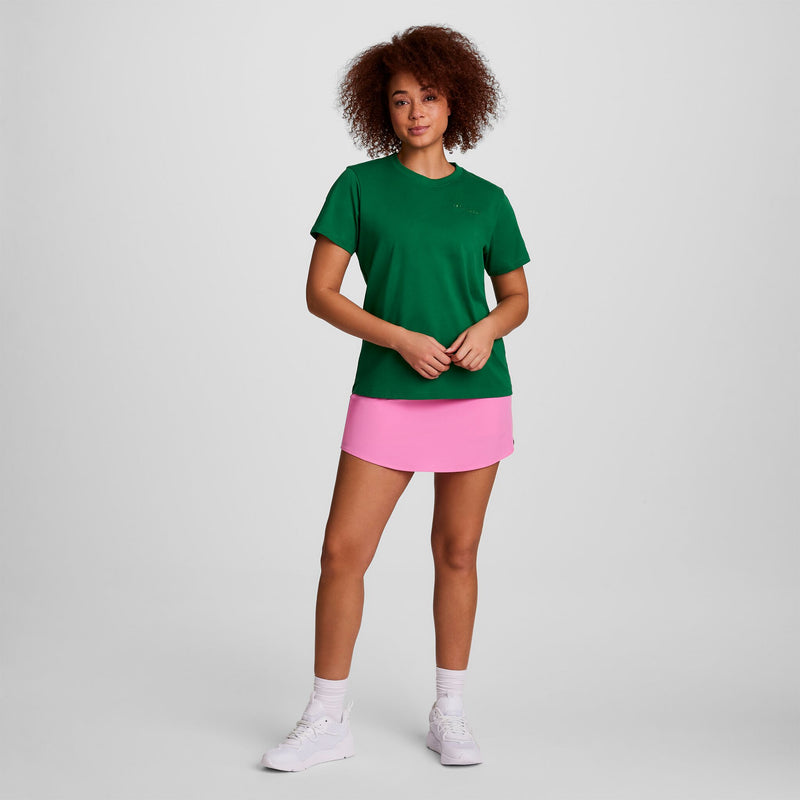 A woman with curly hair wears a green tee, white socks, white sneakers, and the Champion Courtside Skort with C Logo. She stands facing forward against a plain light gray background.