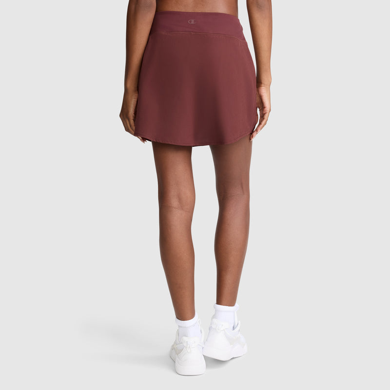 A person is shown from the back wearing the Champion Courtside Skort, C Logo in maroon, paired with white socks and white sneakers, standing against a plain light gray background for a sporty function.