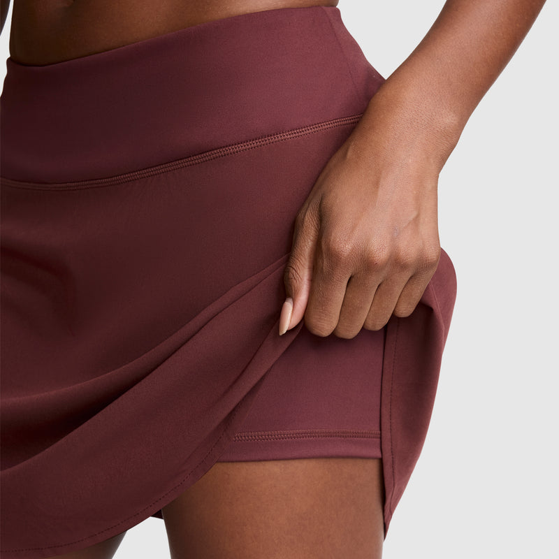 A woman wearing the Champion Courtside Skort, featuring the C Logo in maroon, lifts the hem to reveal matching built-in shorts underneath, emphasizing its sporty functionality. The background is plain and light-colored.