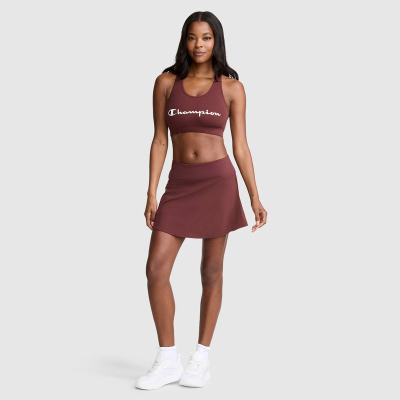 A woman stands against a plain background wearing a maroon Champion sports bra, the Courtside Skort featuring the Champion C logo for sporty function, white socks, and white sneakers. She is smiling with one hand on her hip, embodying effortless court style.