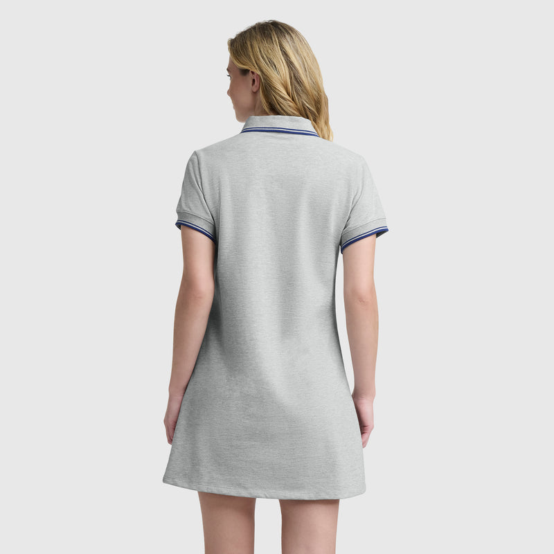 A woman faces away, wearing Champion's Off The Court Polo Dress with the C logo—featuring short sleeves, light gray fabric, and navy-trimmed collar and sleeves—evoking a sleek retro tennis style against a plain white background.