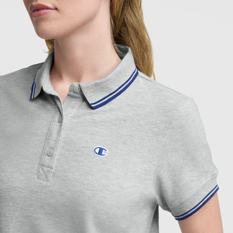 A woman wears the Champion Off The Court Polo Dress in light gray, featuring blue-trimmed collar and sleeves and a small blue C logo on the chest, with light brown hair for a retro tennis-inspired style. Her face is partially visible.