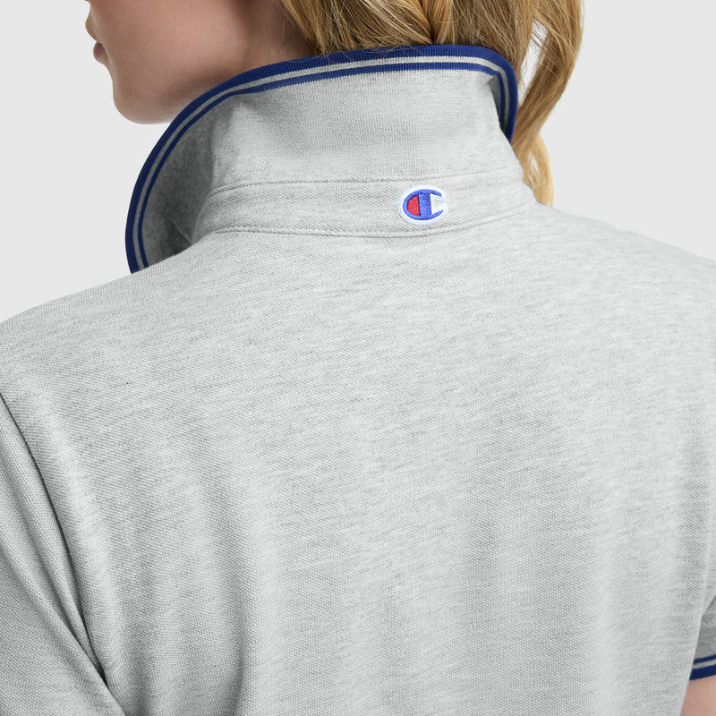 A person with blonde hair is seen from the back wearing the Champion Off The Court Polo Dress, featuring a light gray, high-collared design with blue trim and a C logo on the collar for retro tennis style.