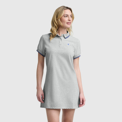 A woman in the Champion Off The Court Polo Dress with C logo, light gray with a collar and dark trim, stands against a plain backdrop, channeling retro tennis style.