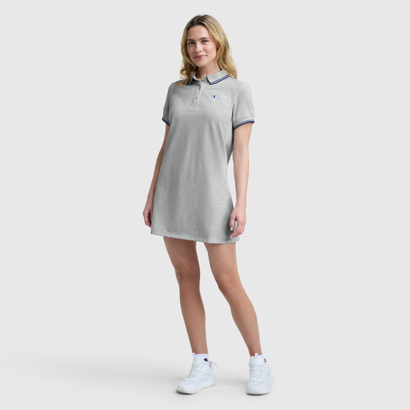 A woman stands against a plain light background, wearing Champion's Off The Court Polo Dress with a C logo. The short-sleeved, light gray dress features navy trim and channels retro tennis style as she smiles softly, holding the hem of her dress.