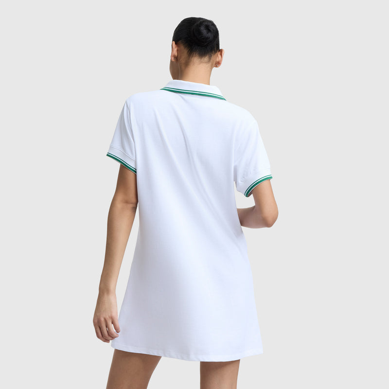 A person stands facing away, wearing the Champion Off The Court Polo Dress with C Logo—a retro tennis-style, short-sleeved white dress featuring green trim and lightweight pique fabric—set against a plain light background.
