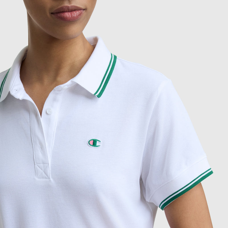 A person wears the Champion Off The Court Polo Dress featuring a green-striped collar and sleeves, lightweight piqué fabric, and a small green and blue C logo embroidered on the chest. The background is plain light gray.
