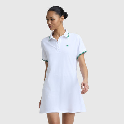 A woman stands against a plain light gray background, wearing the Champion Off The Court Polo Dress with a C logo, featuring retro tennis style, short sleeves, white fabric, and green trim on the collar and sleeves.