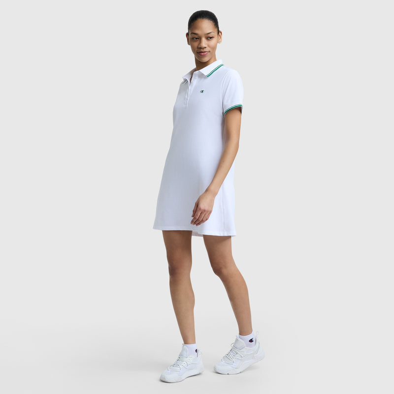 A woman stands confidently in the Champion Off The Court Polo Dress, featuring a C logo and green trim, crafted from lightweight pique fabric, paired with white sneakers and her hair tied back against a plain light gray background.