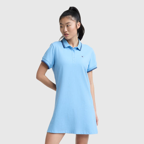 A woman wears the Champion Off The Court Polo Dress, C Logo, in light blue with dark blue trim and retro tennis style. Its short sleeves, collar, and lightweight pique fabric add a sporty yet classic touch against a plain light gray background.
