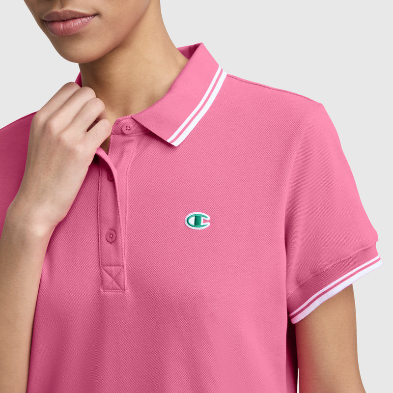 A person stands against a light gray background wearing the Champion Off The Court Polo Dress, featuring white trim and a green and white C logo on the chest for a retro tennis-inspired look.