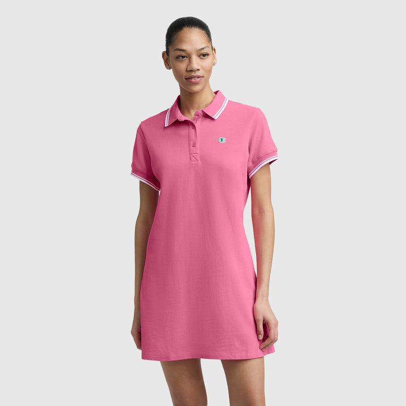 A woman poses against a light gray background wearing the Champion Off The Court Polo Dress, featuring short sleeves, pink fabric, white trim, and a small C logo for a retro tennis-inspired look. Her arms rest relaxed by her sides.