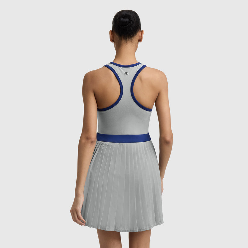 A woman, seen from the back with her hair in a bun, wears the Champion Match Point Tennis Dress featuring a sleeveless grey design with blue trim, pleated skirt, built-in shorts, and an embroidered script logo against a plain background.