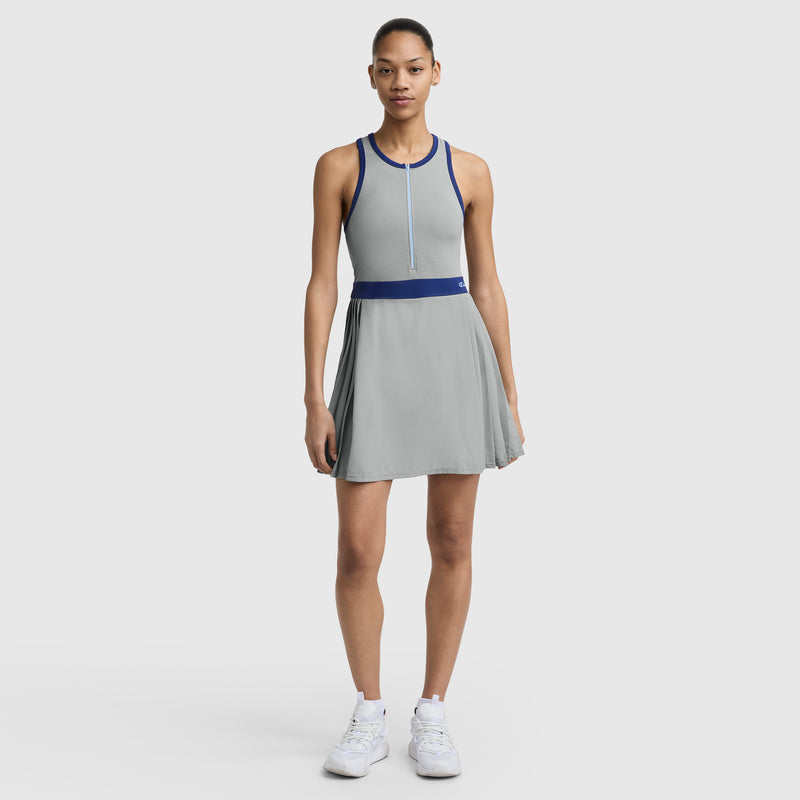 A woman faces forward in the Champion Match Point Tennis Dress with an embroidered script logo, featuring a sleeveless light gray design, blue trim, pleated skirt, built-in inner short, and white sneakers against a plain white background.
