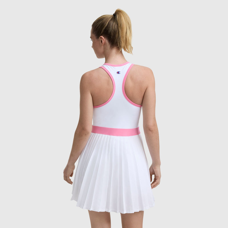 A woman with a blonde ponytail wears the Champion Match Point Tennis Dress with embroidered script logo and pink accents, featuring a pleated skirt, standing with her back to the camera against a light gray background.