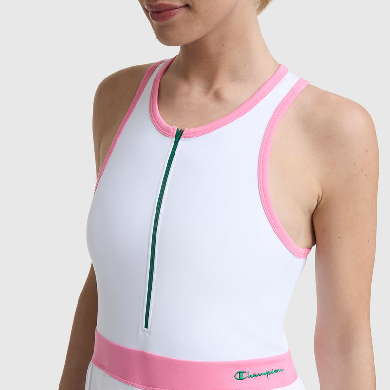 A person wears the Champion Match Point Tennis Dress, featuring a sleeveless white design with pink trim, green front zipper, and an embroidered script logo near the left waist. Image is cropped at the mouth and upper arms.