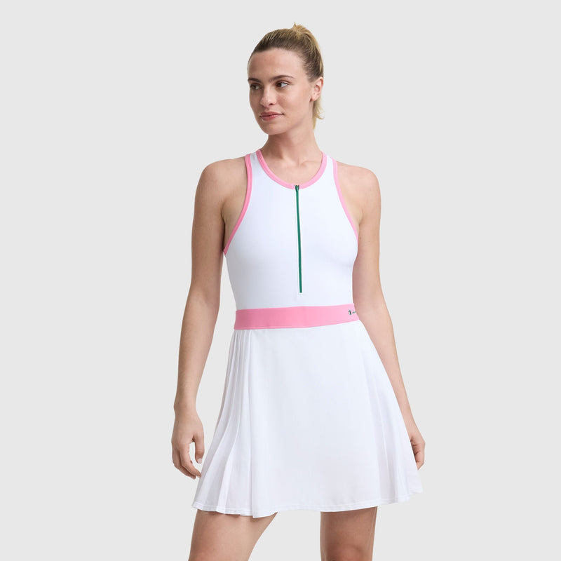 A woman stands against a gray background, looking slightly to the side, wearing the Champion Match Point Tennis Dress with embroidered script logo—a white sleeveless dress featuring pink trim and a green front zipper.