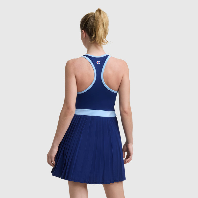 A woman faces away, modeling the Champion Match Point Tennis Dress in navy and light blue with a racerback, pleated skirt, and embroidered script logo, made from recycled polyester, against a plain light background.